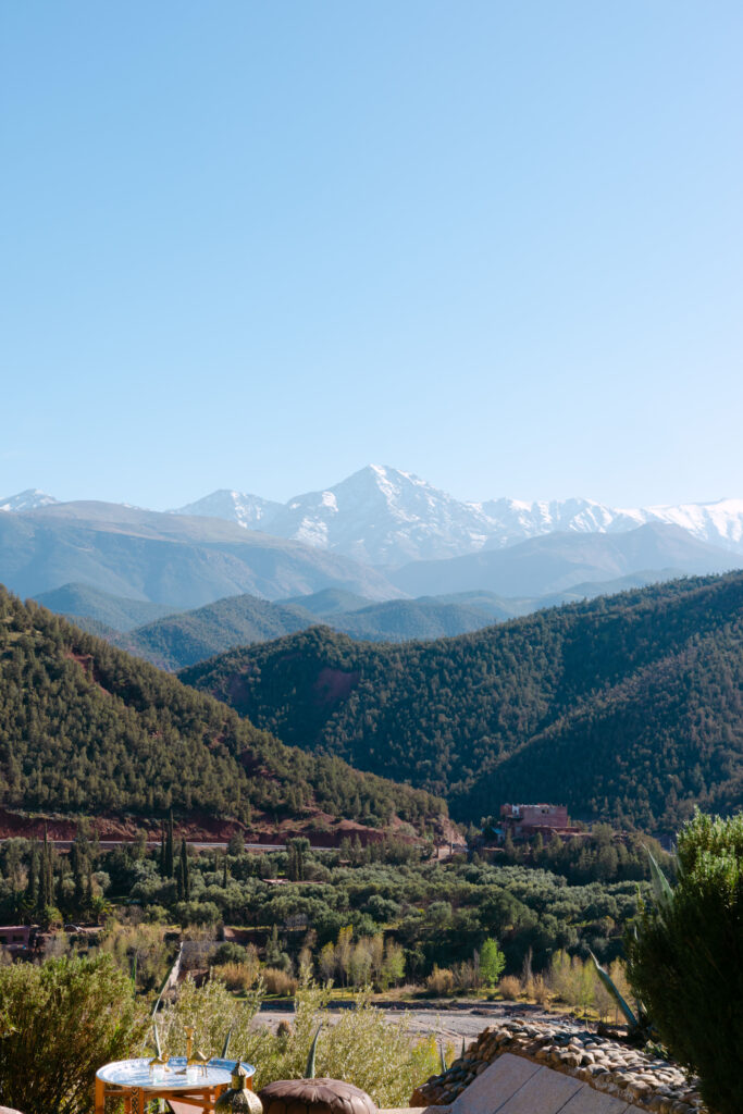 A scenic view of green hills and valleys with snow-capped mountains in the distance under a clear blue sky. A small table with Moroccan tea is visible in the foreground.