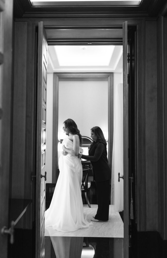 A black and white photo of a bride standing as another woman helps her adjust her dress in an elegant room, viewed through an open doorway.