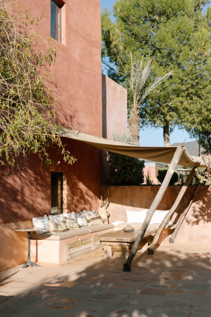 A shaded outdoor seating area with white cushions is set against a terracotta-colored building. A canvas canopy is supported by wooden poles; trees provide additional shade in the sunny courtyard.