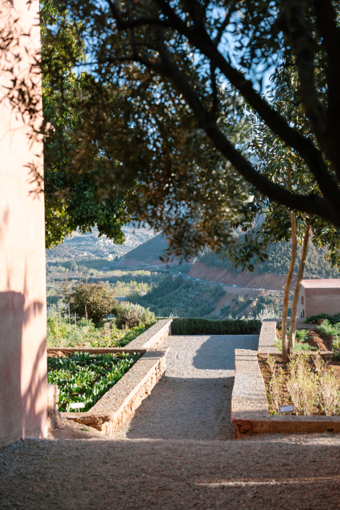 A sunlit garden path bordered by stone walls and greenery leads toward distant tree-covered hills, framed by trees and the corner of a peach-colored building.