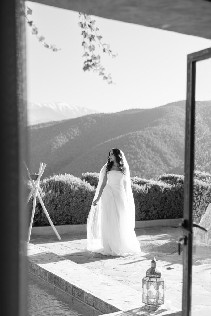 A bride in a flowing white dress and veil stands outside on a stone patio, surrounded by bushes and overlooking scenic mountain views. Sunlight illuminates the scene, and a decorative lantern rests on the ground nearby.