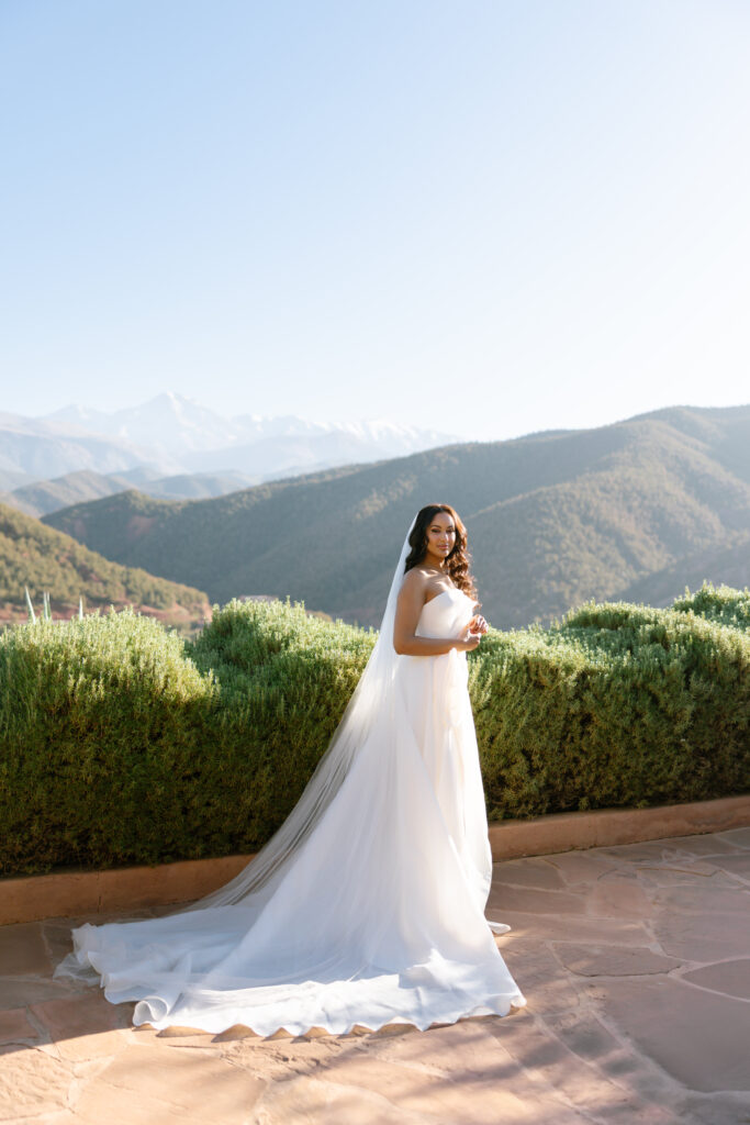A bride in a white wedding dress and veil stands outdoors on a stone patio, surrounded by greenery, with mountains and a clear blue sky in the background.