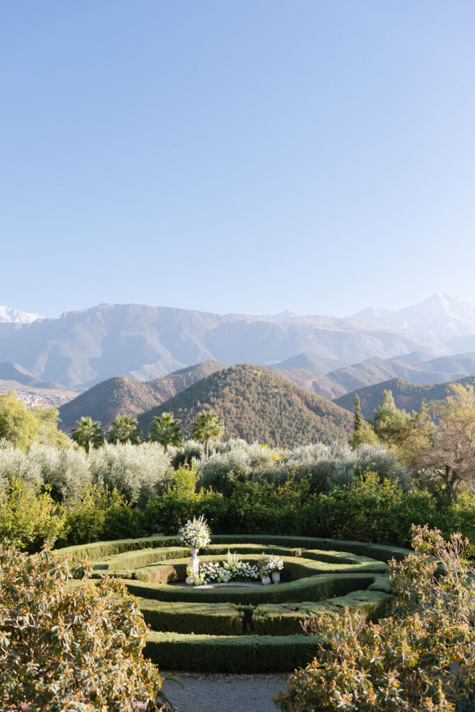 A manicured circular hedge garden with white flower arrangements sits in the foreground, surrounded by lush greenery. Snow-capped mountains rise in the distance beneath a clear blue sky.