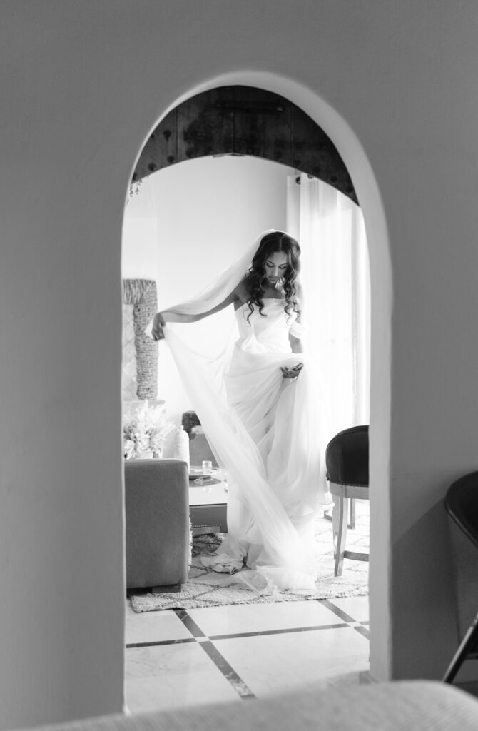 A bride in a flowing wedding dress stands gracefully by a window, holding her veil, seen through an arched doorway in a softly lit room. The image is in black and white.