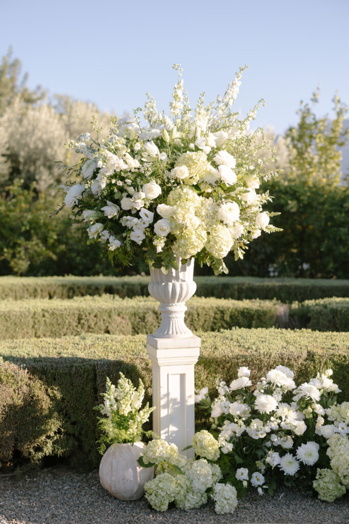 A large white urn holds an arrangement of white flowers and greenery, displayed on a pedestal in a garden. More white flowers and a white pumpkin are arranged at the base, with manicured hedges in the background.