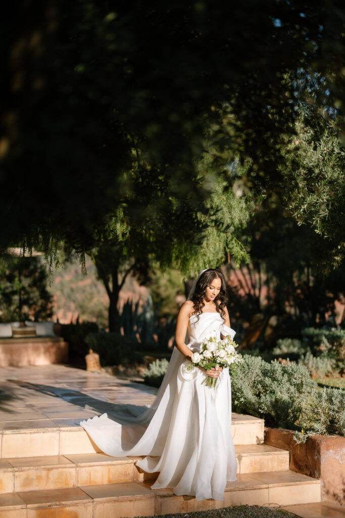 A bride in a flowing white dress walks down sunlit stone steps, holding a bouquet of white flowers, surrounded by lush greenery and dappled sunlight.