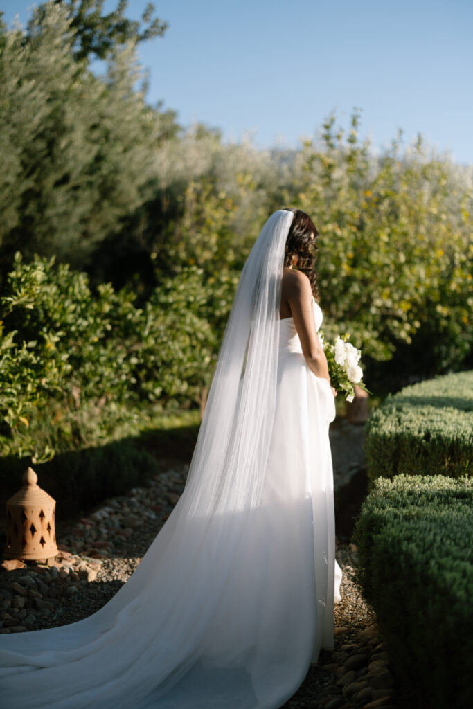 A bride in a long white gown and veil stands in a sunlit garden, holding a bouquet of white flowers. Greenery and manicured hedges surround her as she faces away from the camera.