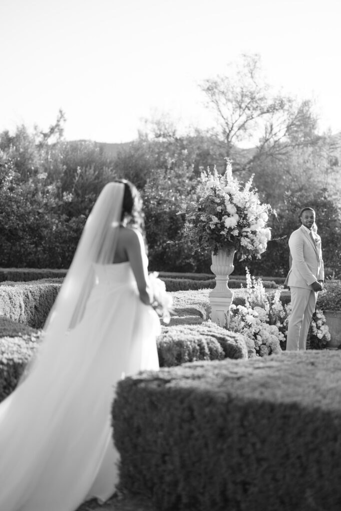 A bride in a flowing gown and veil walks toward a groom standing near a large floral arrangement in an outdoor garden setting, both surrounded by manicured hedges and trees. The image is in black and white.