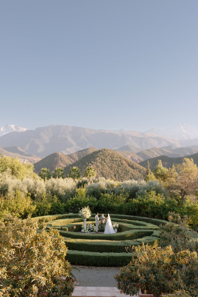 A bride and groom stand in a circular garden surrounded by greenery, exchanging vows with a mountain range in the background under a clear blue sky.