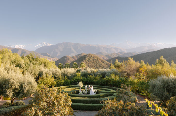 A bride and groom stand in the center of a circular garden hedge, surrounded by lush greenery, with mountains and clear blue sky in the background.