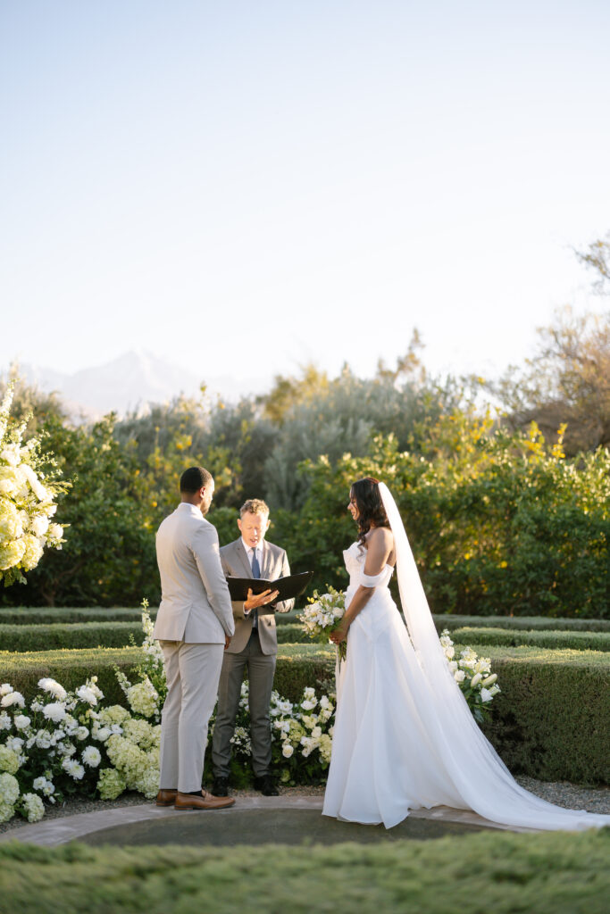 A bride and groom stand facing each other outdoors during their wedding ceremony, with an officiant between them. They are surrounded by greenery and white flowers, under a clear sky.