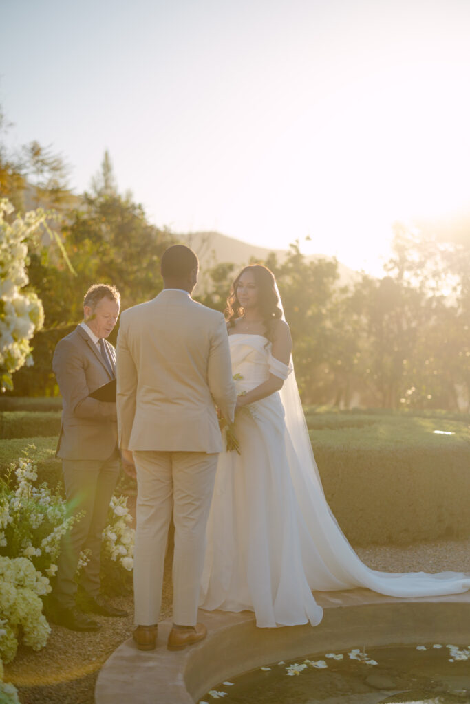 A couple stands facing each other, holding hands during their outdoor wedding ceremony at sunset, with an officiant nearby and lush greenery and white flowers surrounding them.