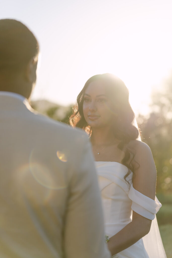 A bride in an off-shoulder white dress stands facing her partner outdoors at sunset, with sunlight creating a warm, glowing effect around her.