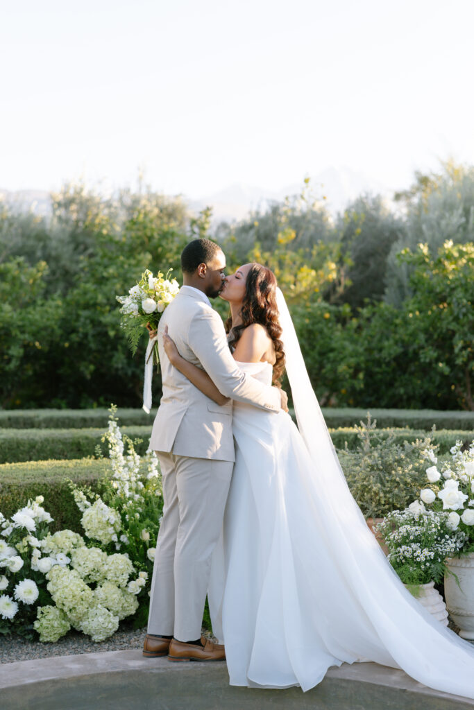 A bride and groom kiss outdoors in a lush garden. The bride wears a strapless white gown with a long veil, while the groom wears a light beige suit and holds a bouquet of white flowers. Greenery surrounds them.