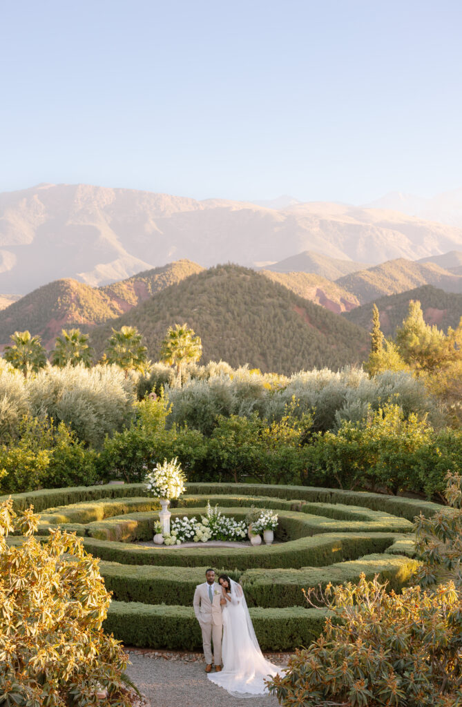 A bride and groom stand together in the center of a manicured garden with circular hedges, surrounded by lush greenery and flowers, with mountains and a clear sky in the background.