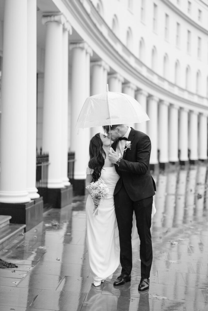 A newlywed couple shares a kiss under a clear umbrella on a rainy day, standing on a wet sidewalk in front of a curved building with tall white columns. The bride holds a bouquet and wears a long white dress.