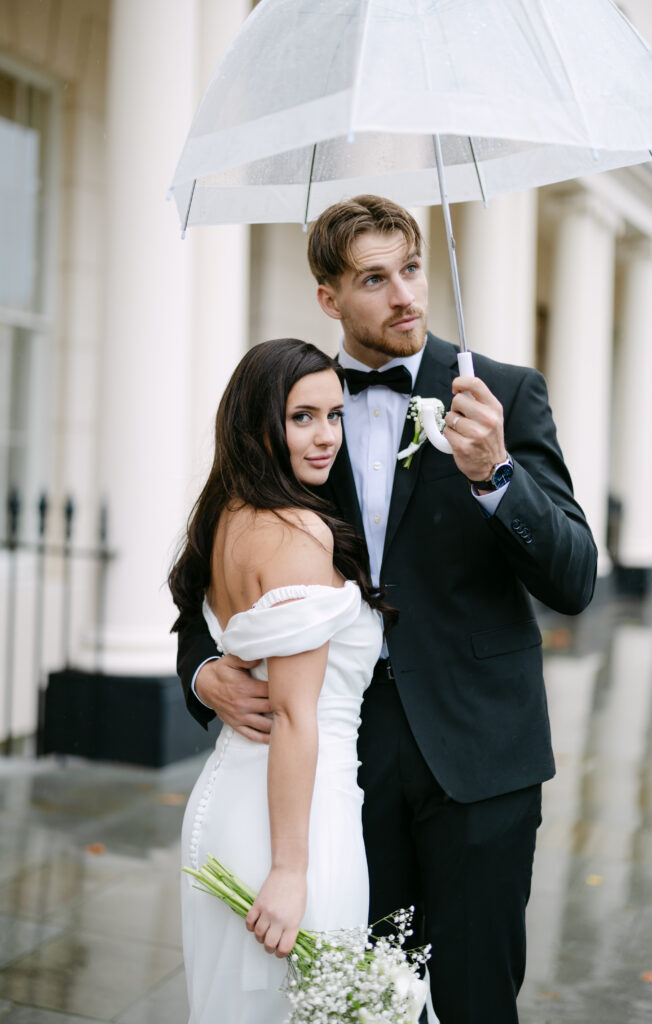 A bride and groom stand outside on a rainy day; the groom holds a clear umbrella over them while embracing the bride, who holds a bouquet and looks at the camera. They are dressed in wedding attire.