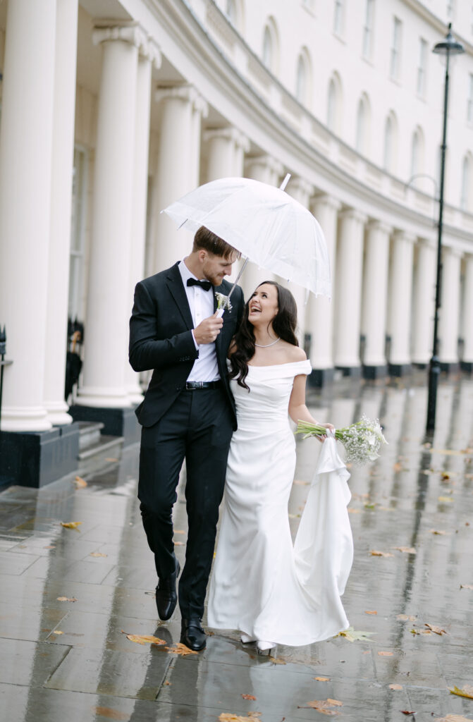 A bride and groom walk arm in arm on a rainy day. The groom holds a clear umbrella over them as they smile and laugh together. The bride wears a white gown, the groom a black suit, and wet pavement reflects their surroundings.