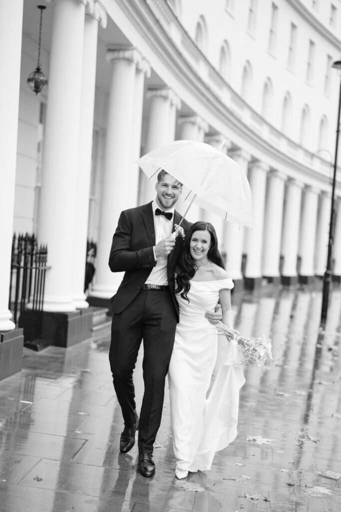 A smiling bride and groom walk arm in arm under a clear umbrella on a rainy day, with elegant white columns in the background. The street and pavement are wet, reflecting the couple and surroundings.