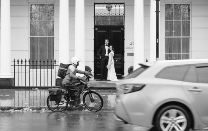 A bride and groom stand on steps in front of a building as a delivery person on a motorbike and a car pass by on a rainy street. The image is black and white.