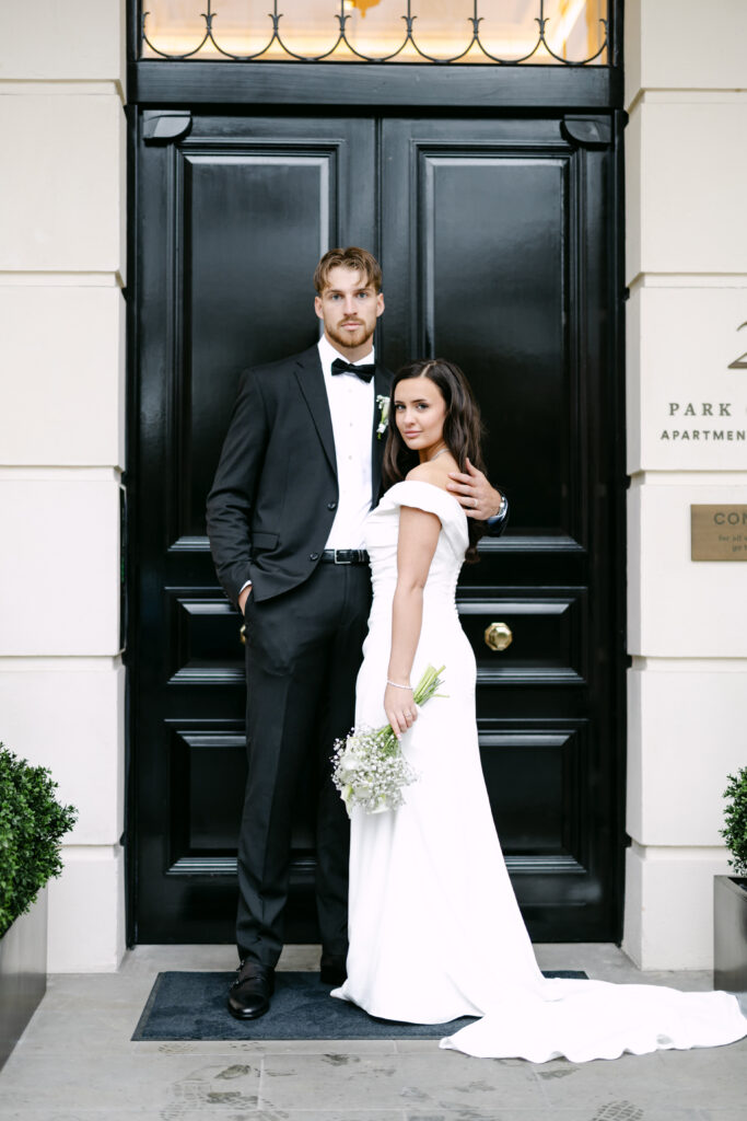 A bride in a white off-shoulder gown holds a bouquet and stands beside a groom in a black tuxedo. They pose in front of a large black door with a brass handle, looking directly at the camera.