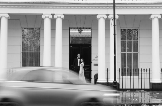A black and white photo of a couple standing in front of a grand building with tall white columns, while a car passes by in the foreground, appearing blurred due to motion.