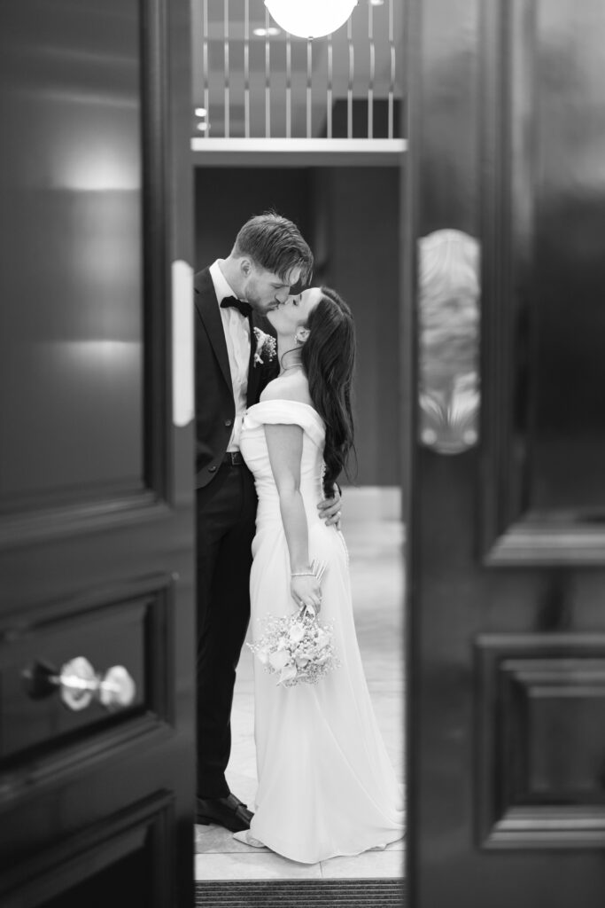 A bride and groom share a kiss indoors. The bride holds a small bouquet and wears a long white dress, while the groom wears a suit. They are framed between an open doorway in a softly lit setting.