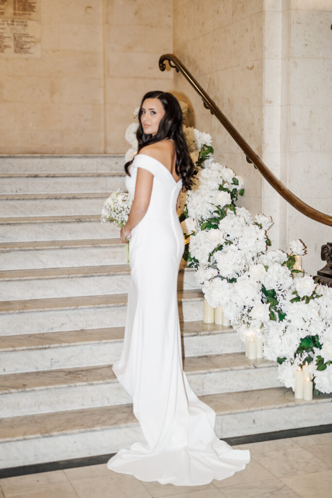 A bride in an off-the-shoulder white gown stands on marble stairs adorned with white flowers and candles, holding a bouquet and looking over her shoulder.