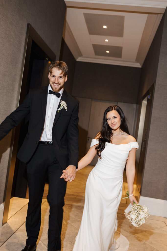 A smiling bride in an off-shoulder white dress holding a bouquet and a groom in a black tuxedo hold hands while walking indoors, looking happy and relaxed.