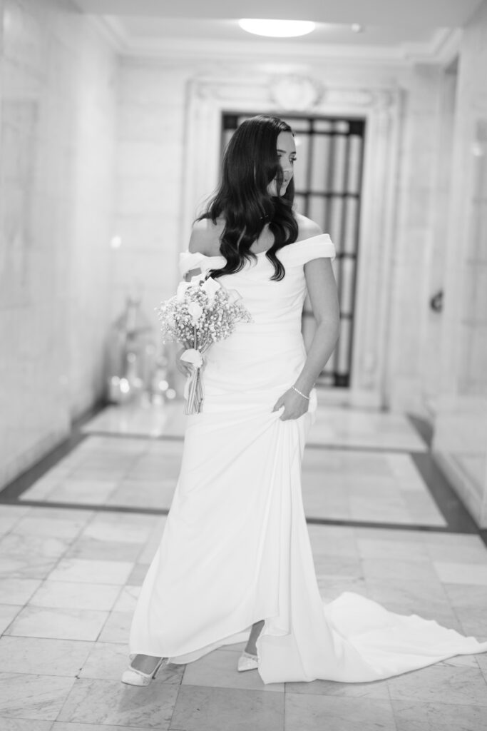 A woman in an off-the-shoulder white wedding gown holds a bouquet and looks to the side while standing in a marble hallway. The image is black and white, with soft lighting and an elegant atmosphere.