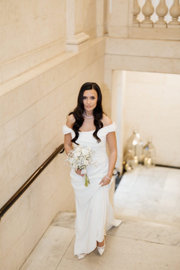 A woman in an off-the-shoulder white wedding dress holds a bouquet of white flowers as she stands on a marble staircase, looking at the camera. The setting is elegant with cream-colored stone walls.