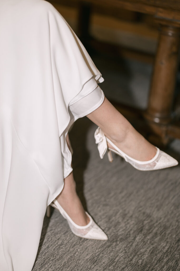 A person wearing a long white dress sits with legs crossed, revealing elegant white high-heeled shoes with bows. The scene appears indoors on a gray carpet next to a wooden table.