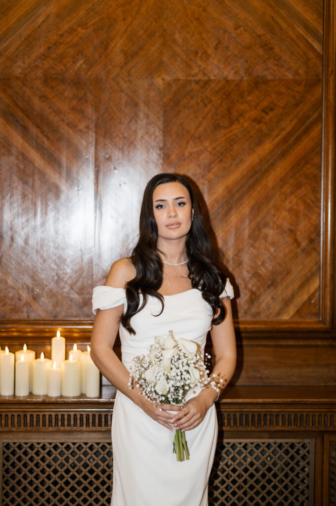 A woman in an off-shoulder white dress holds a bouquet of white flowers, standing indoors in front of a wooden wall with lit candles to her left.