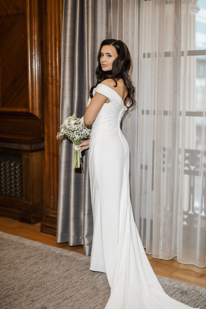 A woman in an elegant off-the-shoulder white wedding dress stands indoors, holding a bouquet and looking over her shoulder toward the camera, with long dark hair cascading down her back.