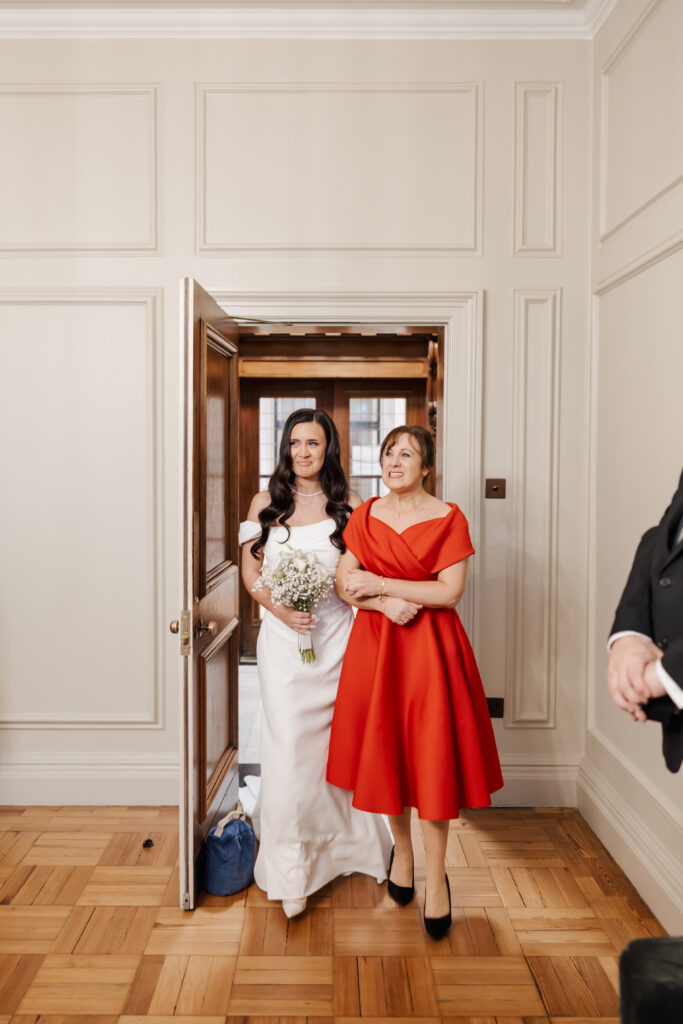 A bride in a white dress holding a bouquet stands arm-in-arm with a woman in a red dress, walking through a doorway into a bright room with wood floors and cream-colored walls.