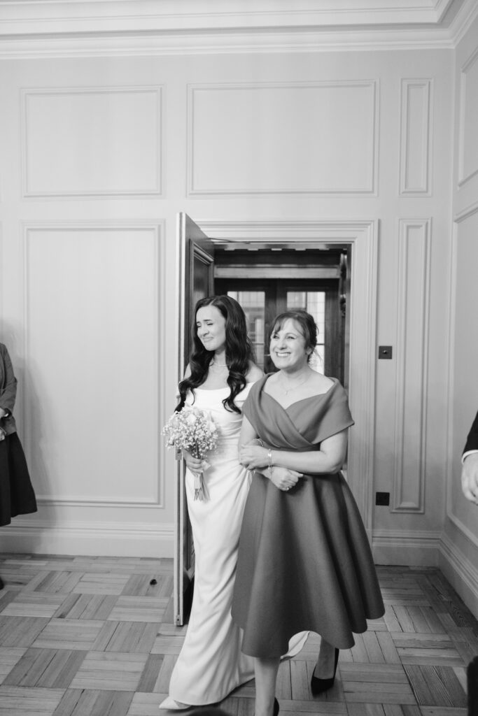 A bride in a white dress holding a bouquet walks arm-in-arm with a woman in a knee-length dress, both smiling, through a doorway into a room with paneled walls. The scene is in black and white.