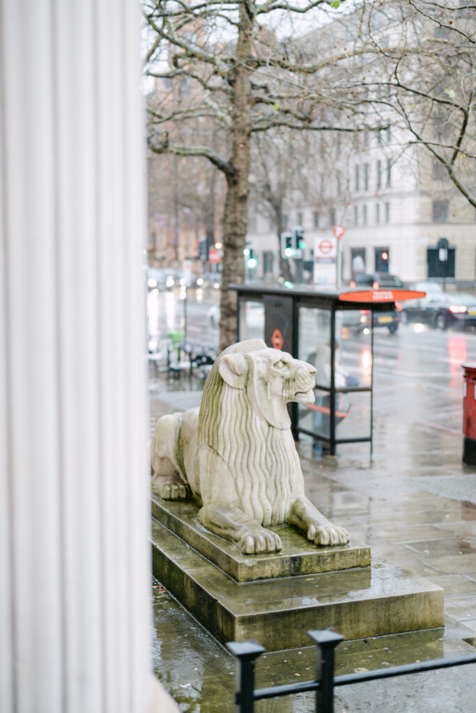 A stone lion statue sits on a pedestal beside a wet city sidewalk on a rainy day, with a bus stop, traffic lights, and cars in the background. Bare trees line the street.