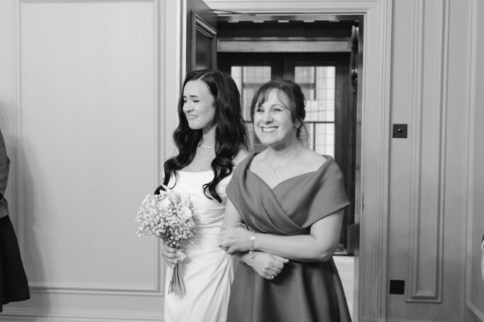 A bride in a white dress holding a bouquet walks arm-in-arm with a woman in an off-the-shoulder dress. Both are smiling, and the scene appears to be indoors at a formal event, possibly a wedding.