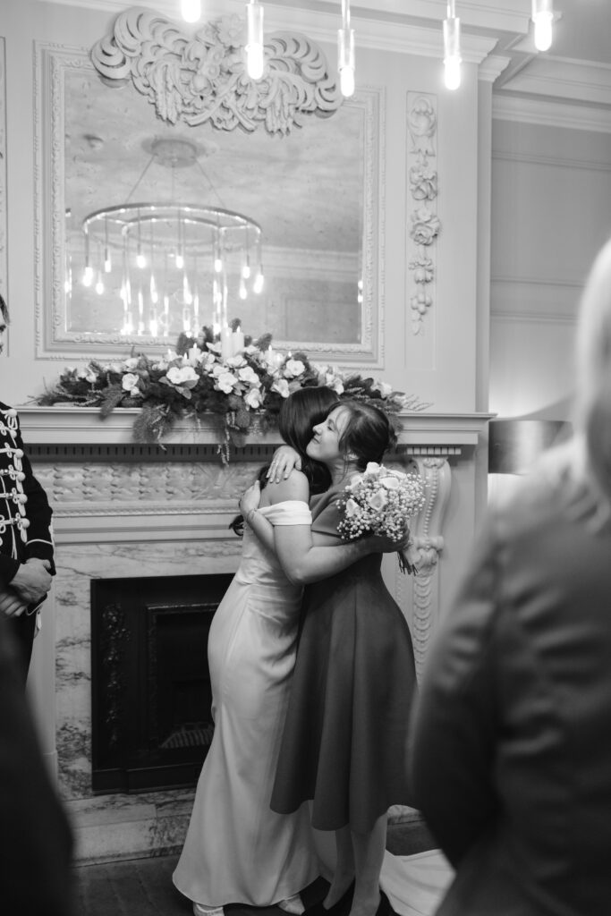 Two women embrace in front of a decorated fireplace, one wearing a long white dress and the other holding a bouquet of flowers. The scene appears emotional, with onlookers nearby. The setting is elegant and warmly lit.