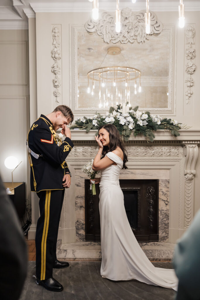 A bride in a white gown and a groom in a military uniform stand in front of a decorated fireplace, both wiping away tears with smiles during an emotional wedding ceremony.