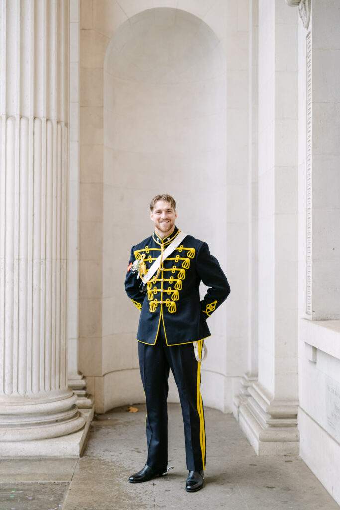 A man dressed in a formal navy blue military uniform with gold trim stands smiling in front of tall white columns and an arched stone wall.