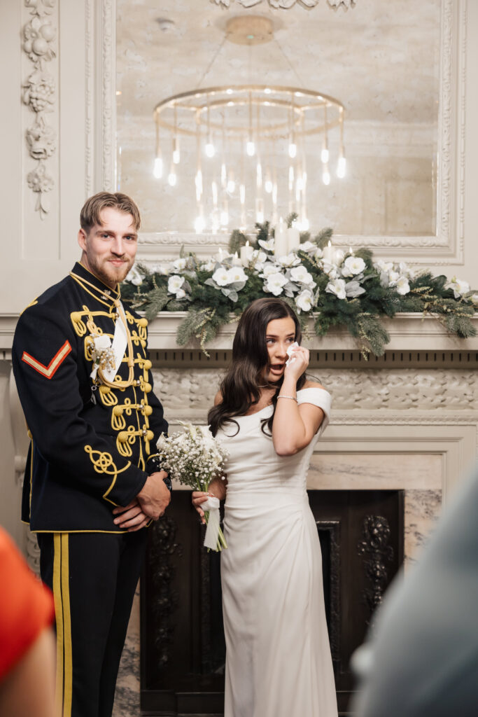 A bride in a white dress wipes a tear from her face while holding a bouquet, standing next to a groom in a decorative military uniform. They are indoors in front of a decorated mantel with white flowers and greenery.