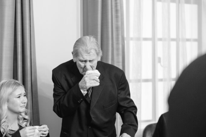 An elderly man in a suit wipes his nose with a tissue, while a young woman beside him appears emotional. Both are indoors near large windows with curtains, and the mood seems somber.