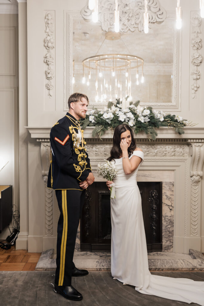 A bride in a white dress holds a bouquet and wipes a tear while standing beside a groom in a formal military uniform in front of an ornate fireplace decorated with white flowers and greenery.