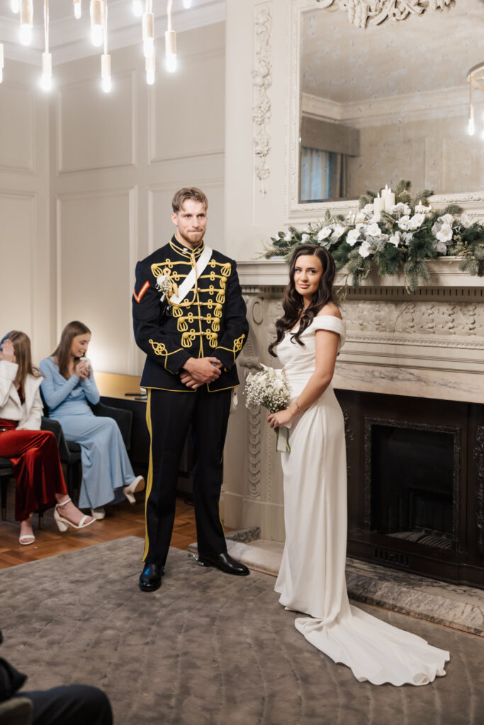 A bride in a white gown and a groom in a formal black and gold military uniform stand together in an elegant room with a marble fireplace and floral arrangements, while guests sit nearby.