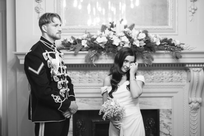 A man in a decorated uniform stands beside a woman in a white dress holding flowers, who wipes her eye. They are in front of an ornate fireplace with a floral arrangement above it.