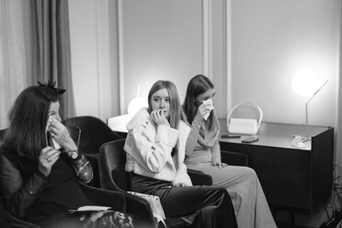 Three women sit in a row, visibly emotional and wiping tears from their eyes. The setting appears to be indoors, with a desk and glowing lamps in the background. The scene is in black and white.