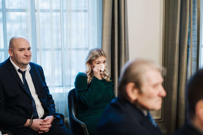 A woman in a green dress wipes her eyes with a tissue, sitting among other people in formal attire in a room with large windows and curtains. The mood appears somber or emotional.