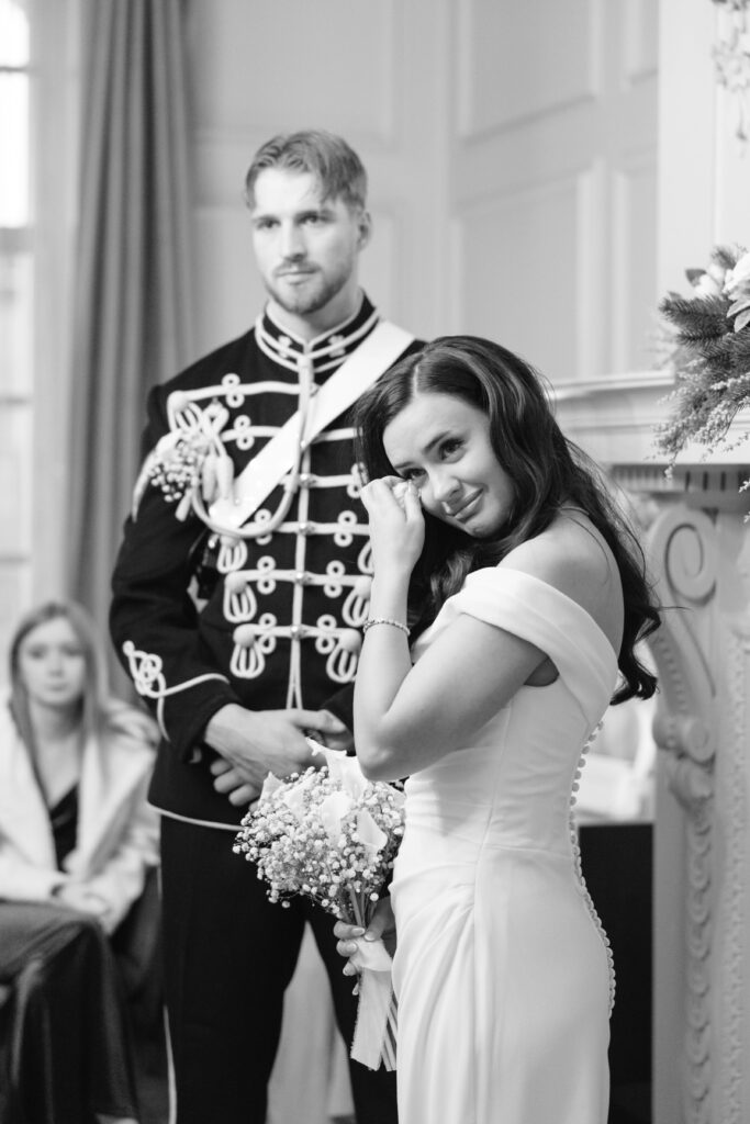 A bride in a white gown wipes a tear while holding a bouquet, standing beside a groom in a decorated military-style uniform. A seated guest is visible in the background. The scene appears emotional and formal.