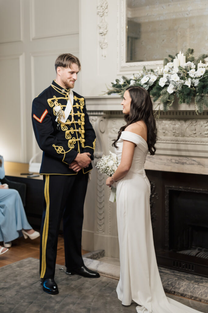 A bride in an off-shoulder white gown and a groom in a formal military uniform stand facing each other during a wedding ceremony in an elegant room with a decorated mantelpiece.
